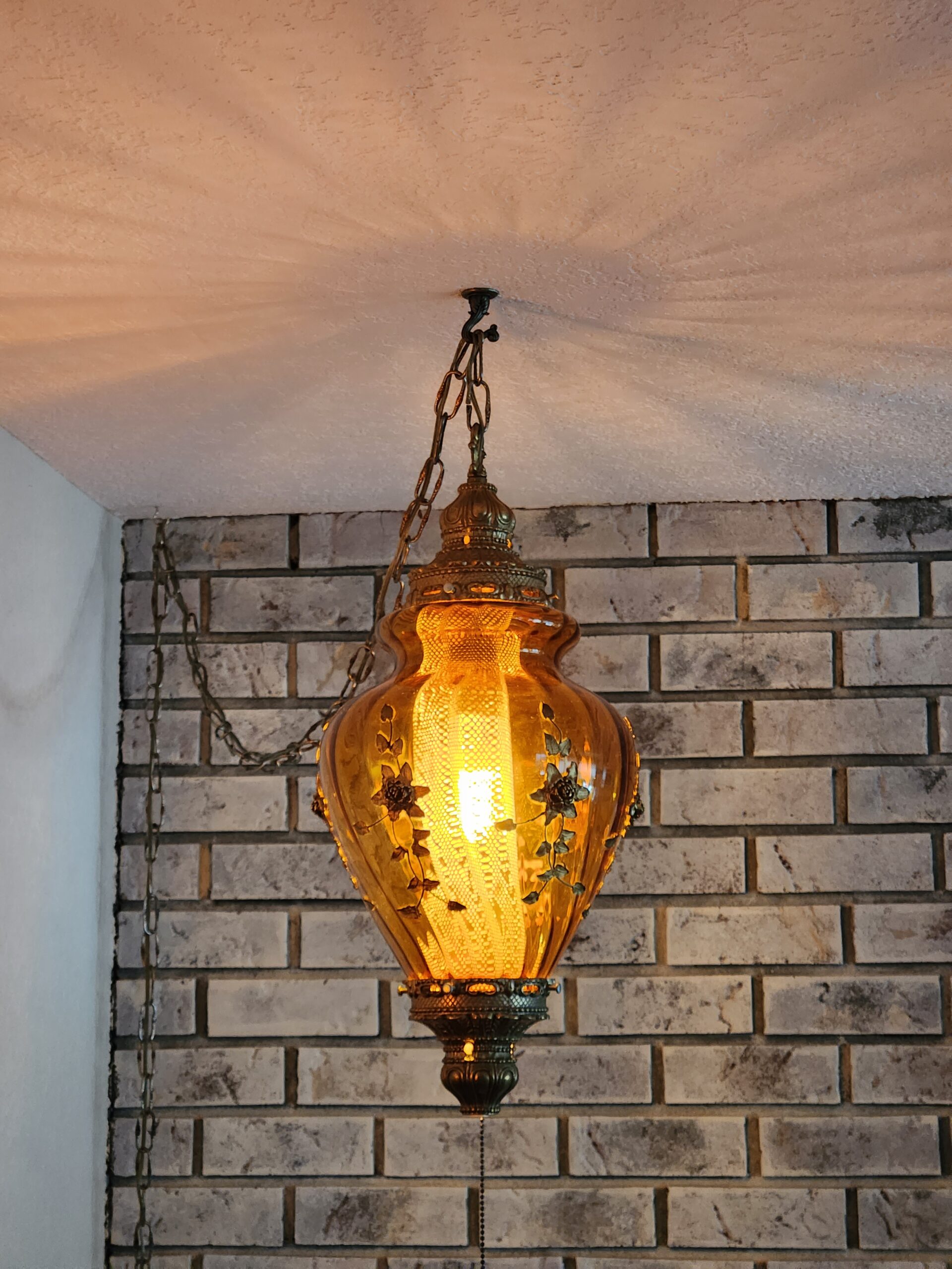 An amber glass vase shaped lamp hangs from the ceiling. It has an ornate brass top and bottom, and golden flowers and vines on the glass. 