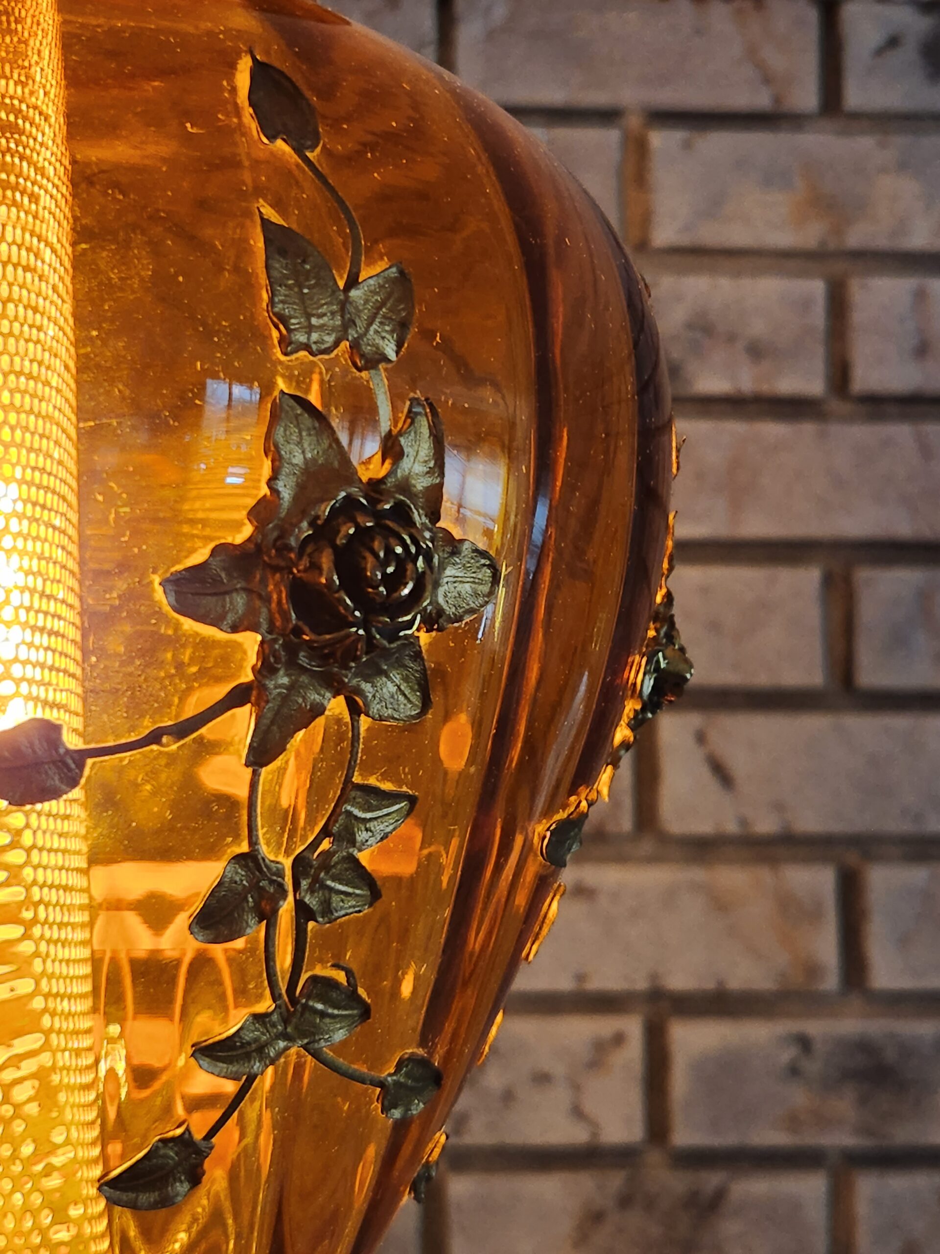 Close up of an amber glass vase shaped lamp with golden flowers and vines on the glass. 
